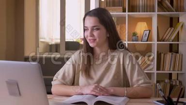 年轻有魅力的白种人女学生在笔记本电脑上阅读一本书并进行视频通话的特写照片